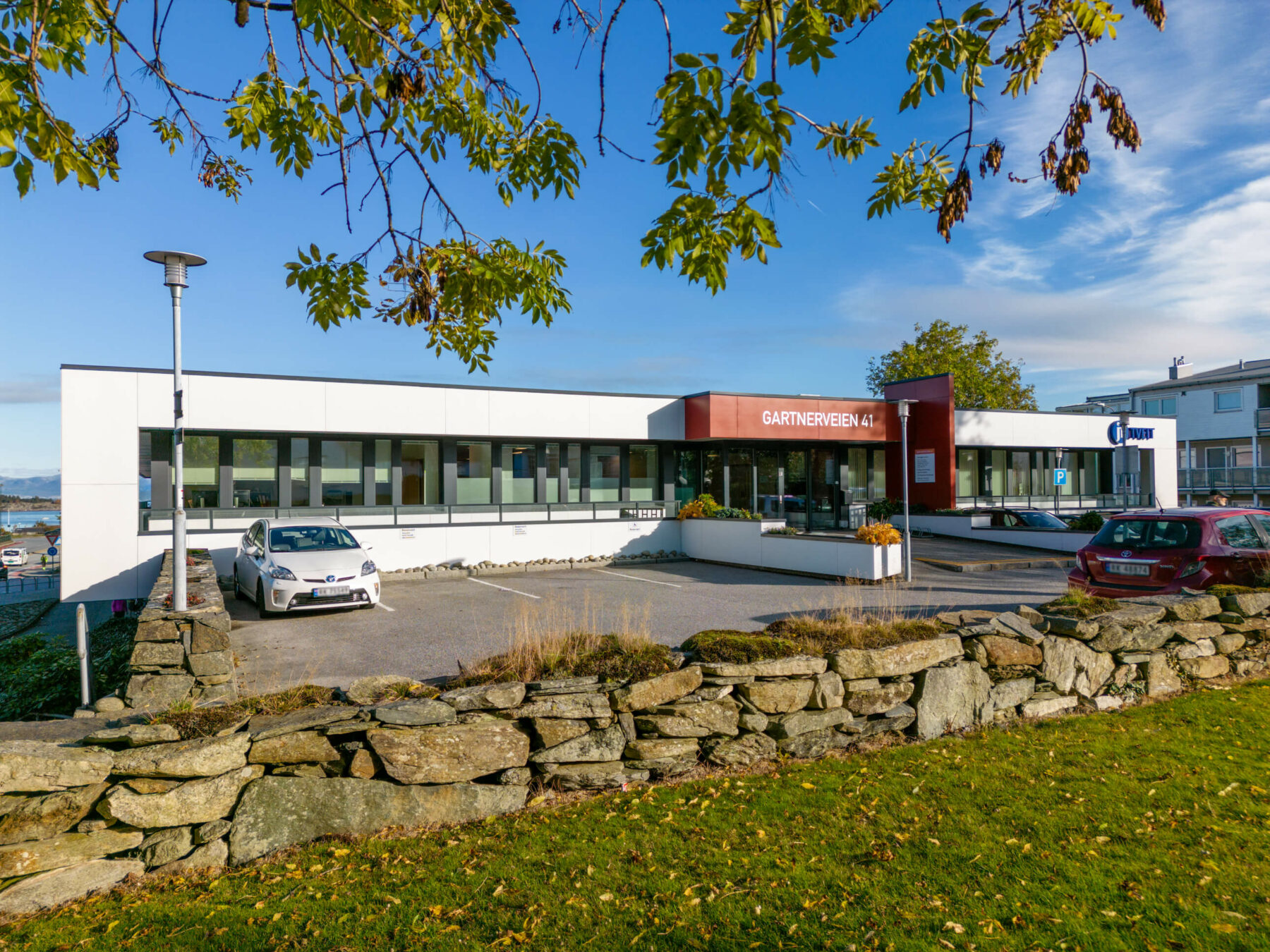 Avlang hvit bygning med parkering foran og ett steingjerde rundt med grønn plen.